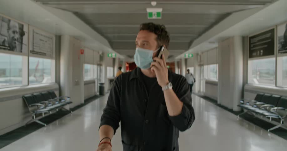 A man walking through an airport wearing a mask answers his phone