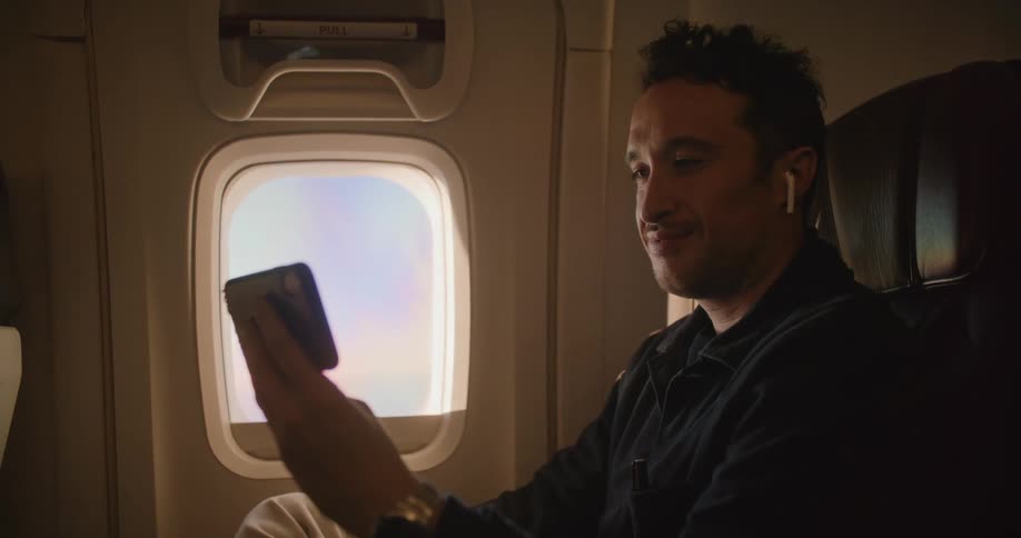 A man wearing wireless earbuds laughes while watching a video on the screen on his smartphone while riding in an airplane