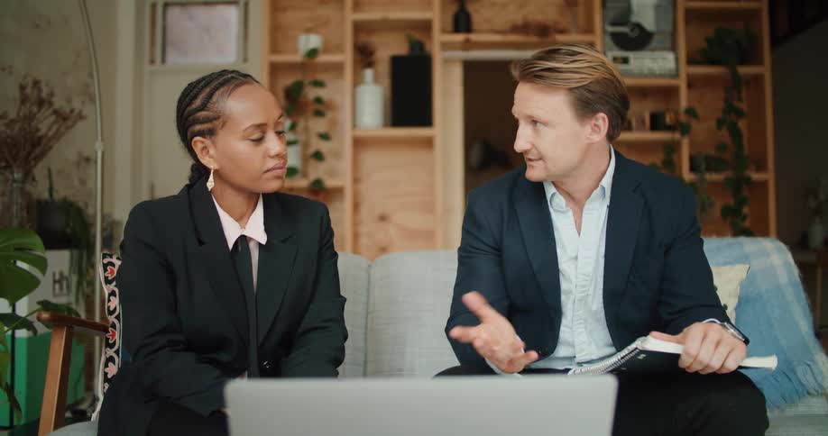 Two coworkers in suits chatting while sitting on a couch in front of a laptop