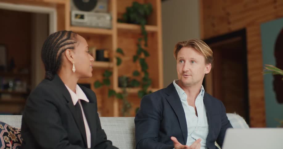 A man and woman in business attire sit side by side on a couch talking