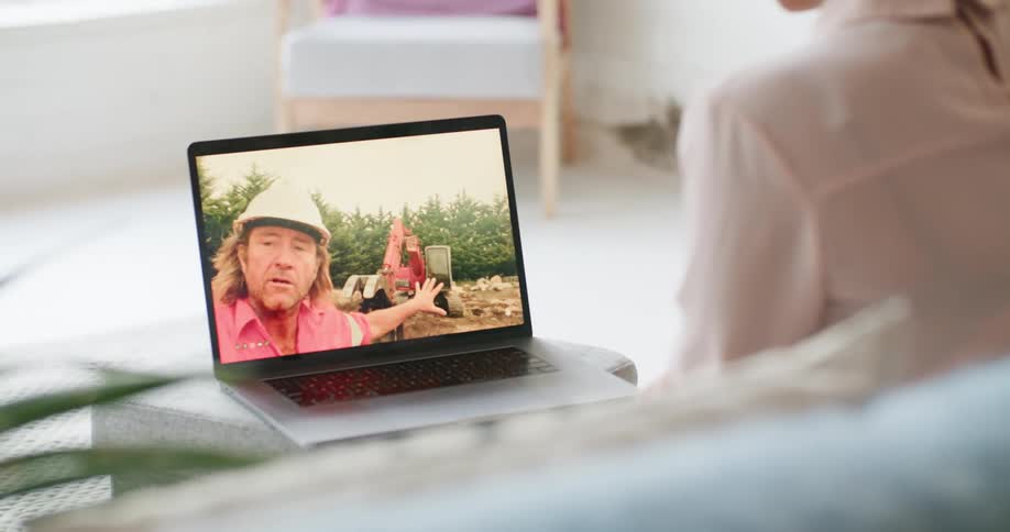 A woman sits at home and watches a constructor worker on a laptop