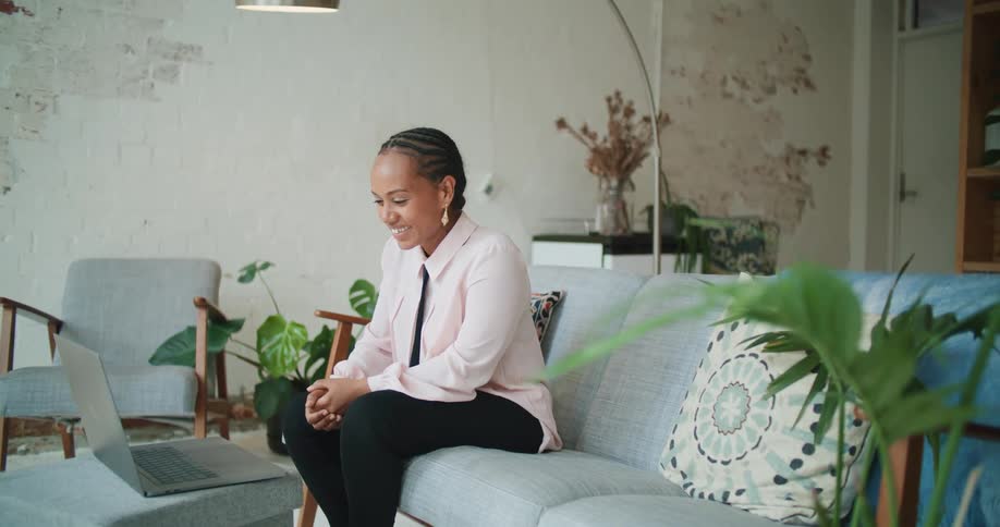 A woman sitting on a sofa in the living room of a house uses a laptop to participate in a video conference