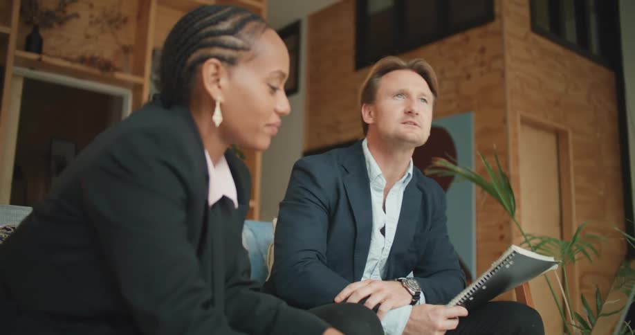A man sitting on a sofa holding a spiral notebook talks with a woman using a laptop while sitting next to him