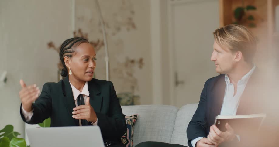 A man nods along while listening to his coworker as they sit on a couch in front of a laptop