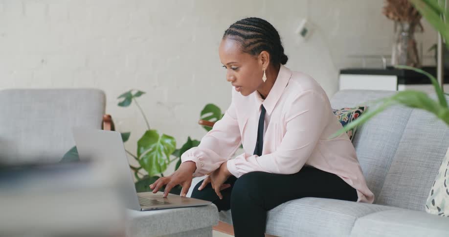A woman sitting on a sofa in the living room of a house during the day uses a laptop to participate in a video conference