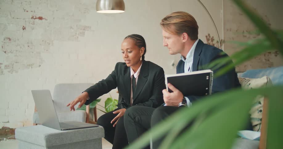 A man and a woman sitting on a couch talking and looking at a laptop with a wall in the background