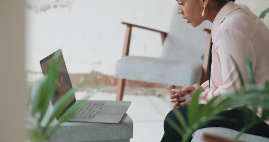 A woman sitting on a sofa in the living room of a house gestures as she participates in a video conference using a laptop