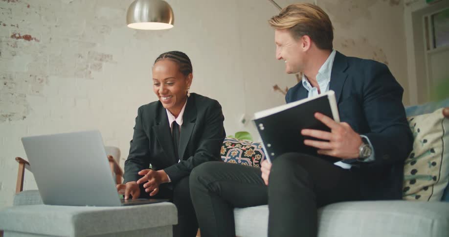 A man holding a notebook sits on a couch with a woman who is using a laptop and they talk and laugh