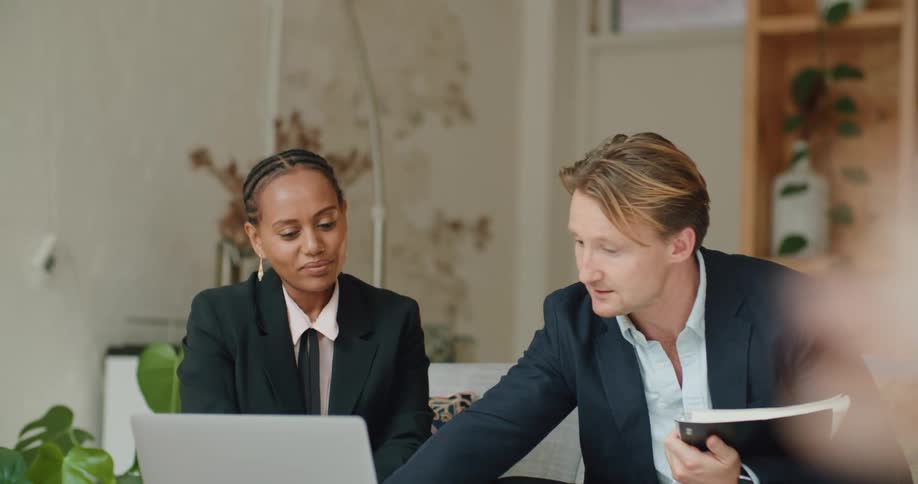 Two coworkers in suits talking while looking at a laptop on a couch