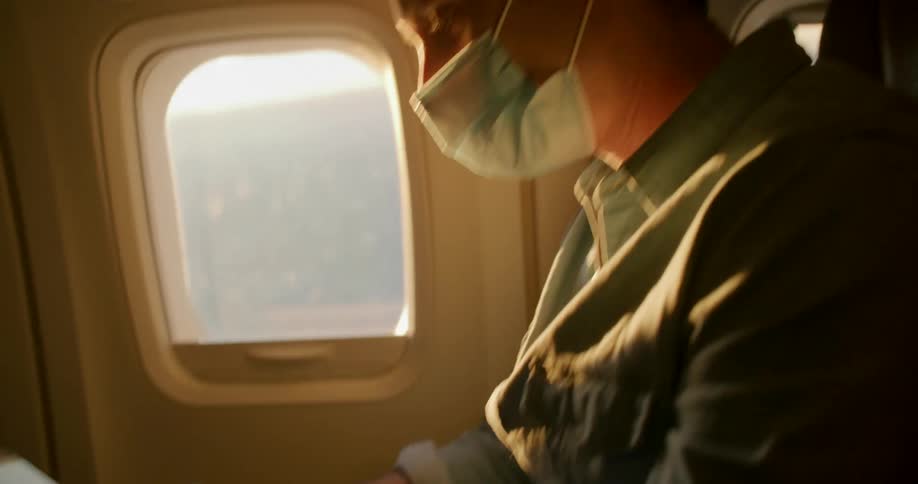 A plane passenger sits in a window seat with a face mask on and texts on his phone