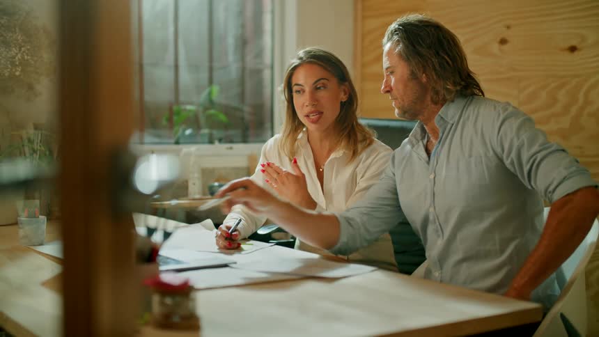 A man and a woman sitting at a desk pointing at a laptop with a window in the background