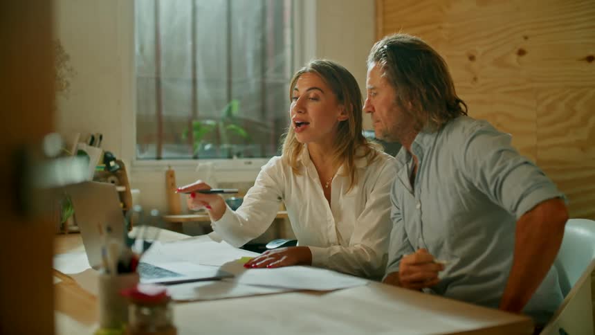 Two coworkers talk and point while sitting a desk in front of a laptop