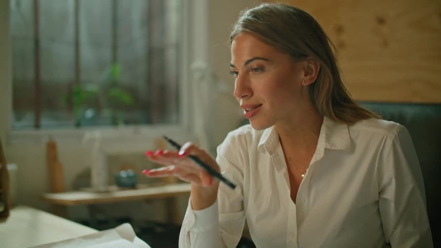 A woman sitting at a desk in an office gestures with her hands as she talks and participates in a video conference with her laptop