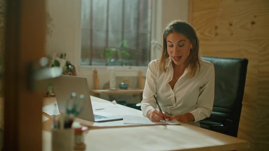 A woman sitting at a desk writing on a paper with a pen while video chatting on a laptop