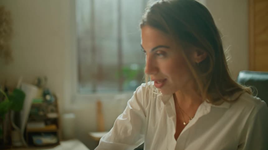 A woman sitting at a desk with a pen in her hand video chatting on a laptop with a window in the background