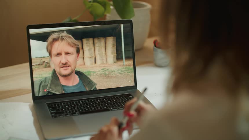 A man stands outdoors while talking to a woman who is sitting indoors at her laptop