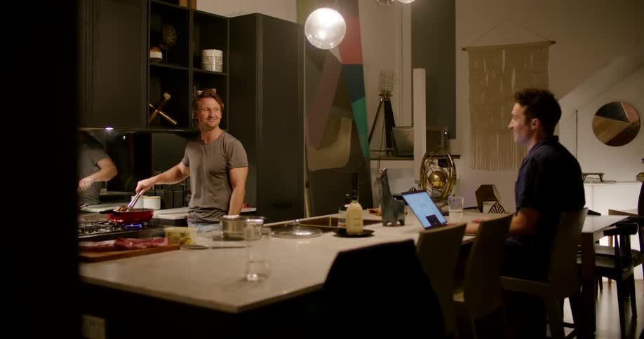 A man sitting in front of a laptop at a kitchen counter talks with a man cooking in a skillet on the other side of the counter