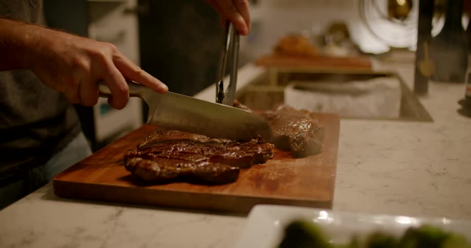 A man chats and laugh as he stands in his kitchen cutting steak on a wooden cutting board