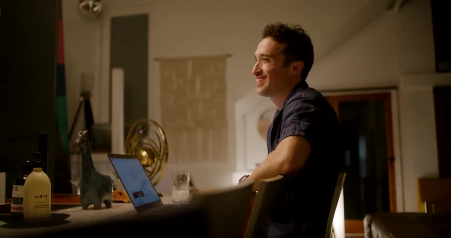 A man laughs and gestures while sitting at his desk in front of a laptop and talking to someone
