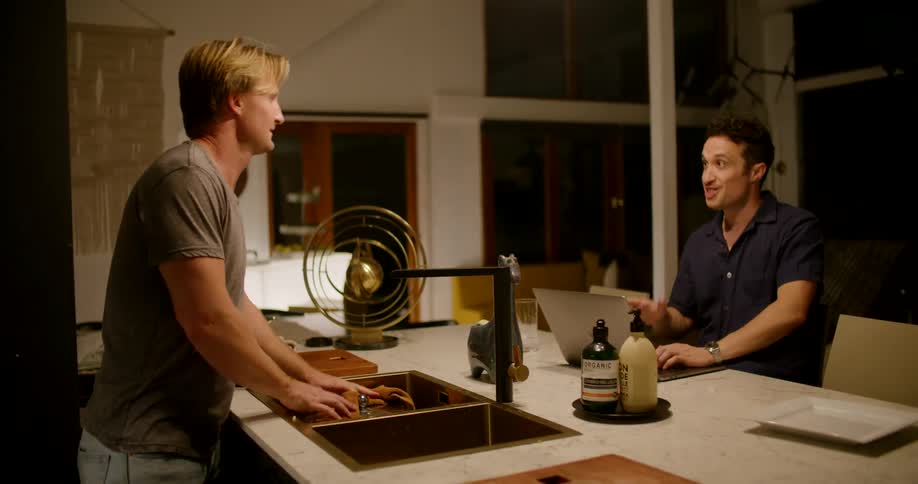 A man at home takes a pot off the stove and sets it in the sink and talks with the man sitting at the counter on his laptop