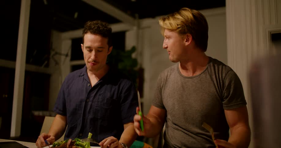 Two men sit at home and eat a meal and talk