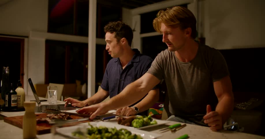 A man listens to his friend talk and eats a piece of steak while plating dinner
