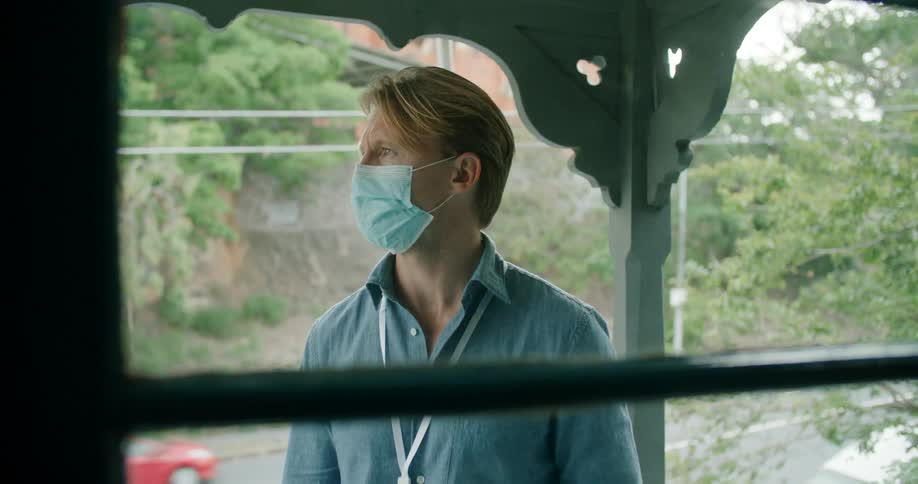A man wearing a face mask stands outside a home holding a notebook as traffic passes by