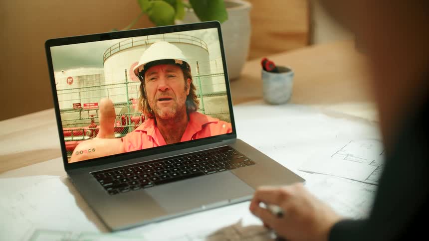 A woman writing at a table returns the wave of a man being displayed on the laptop before her