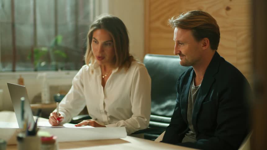 A woman writes on a sheet of paper while speaking with a man sitting beside her at her desk