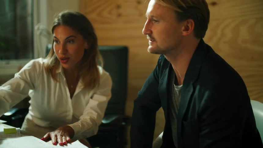 A woman gestures at documents and a laptop while speaking to a man sitting beside her at her desk