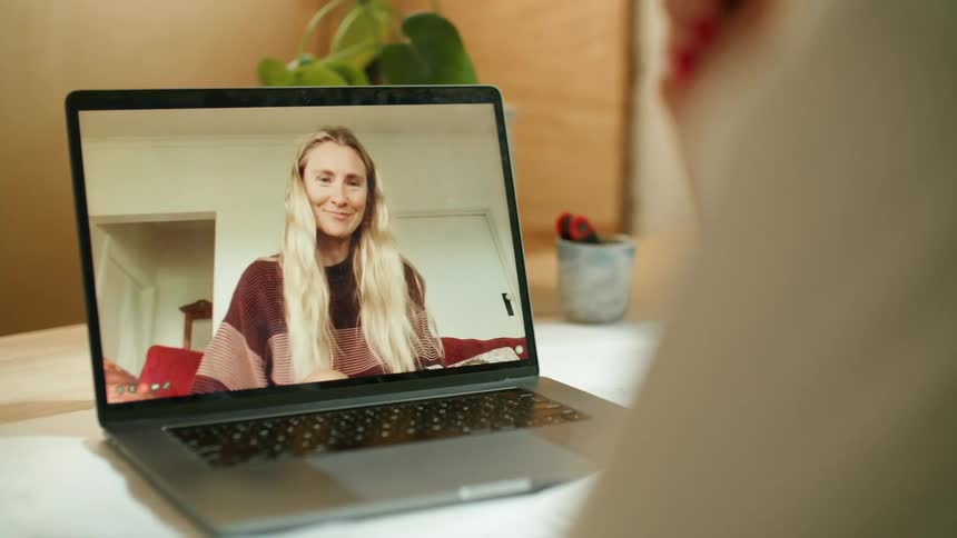A woman in a room sitting at a desk video chatting on a laptop with a plant in the background