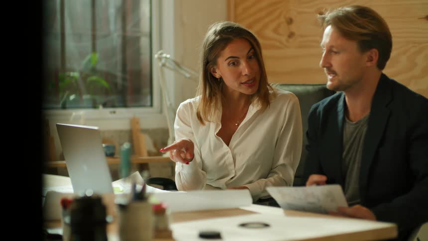 A woman speaks to a man sitting beside her at a desk as he nods along to her words while holding a sheet of paper