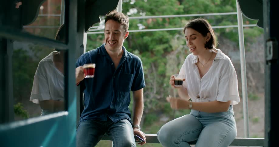 A man and woman drink and hold a conversation while sitting beside each other on the railing of a balcony over a road during the day