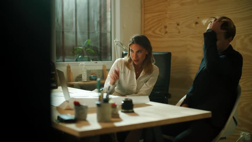 A man and a woman sitting at a desk pointing at a laptop with a window in the background