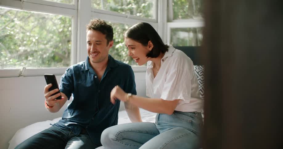 A woman waves to someone on a smartphone being held by her partner as they sit on the edge of a bed in a small cottage