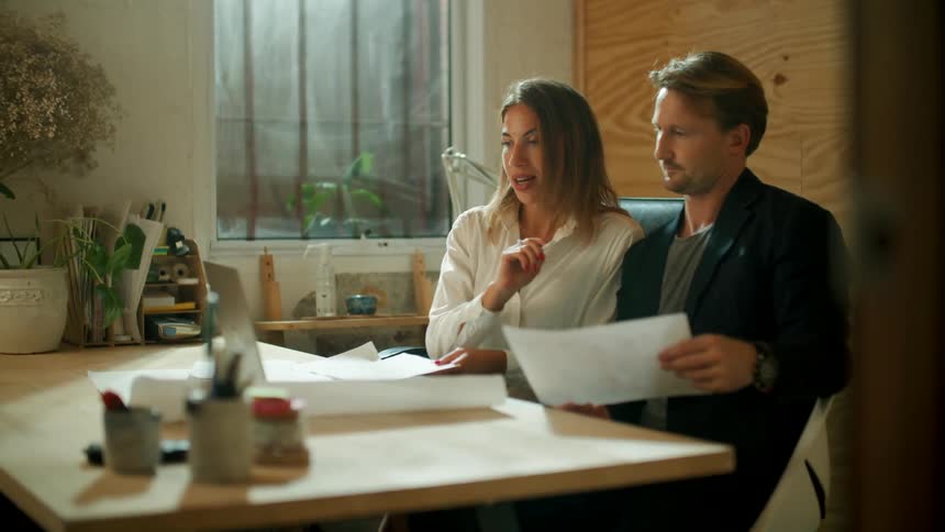 A man and woman sit in an office and review some documents while video conferencing with someone via a laptop