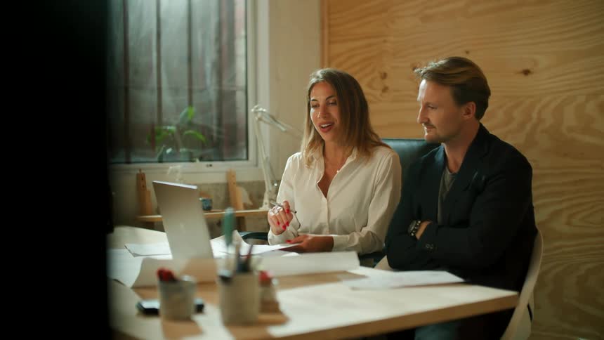 Two coworkers laugh and sit side by side as they talk to someone via video call on a laptop