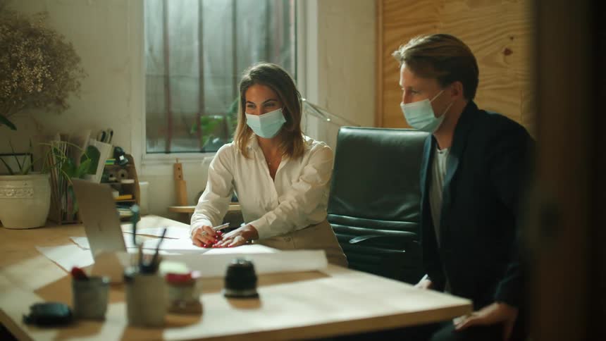 Two coworkers sit apart and wear face masks as they wave at someone during a video call on a laptop
