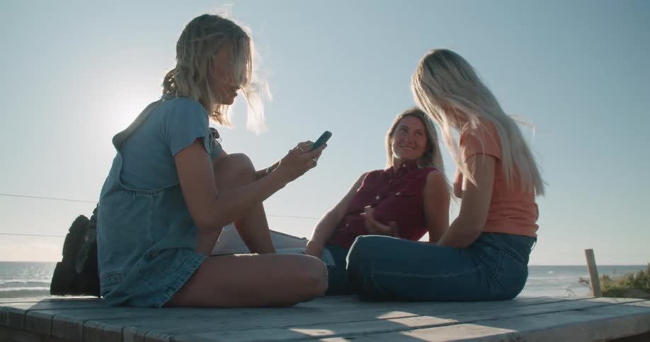 Three teenage girls sit on the shore and talk and laugh as one of them scrolls on her cell phone