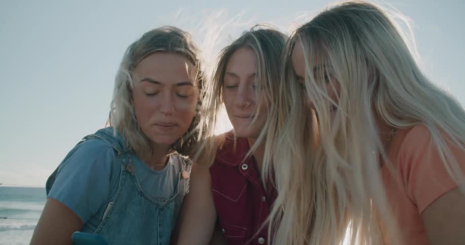 Three blonde women sit on a wooden dock near the ocean talking on one phone as the wind blows their hair