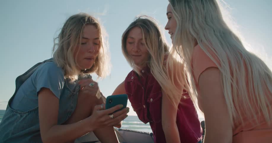 Three teenage girls sit on the shore and talk and look at a cell phone