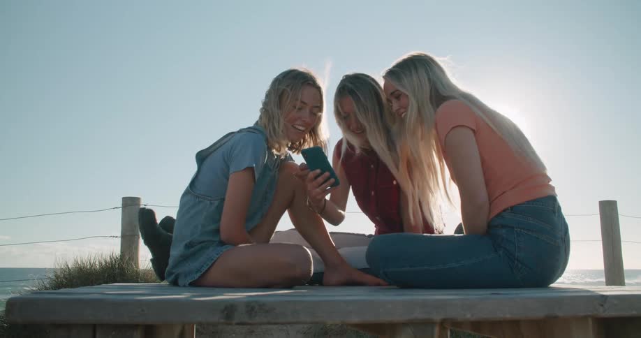 Three girls sit around outside and are laughing while looking at one girls phone