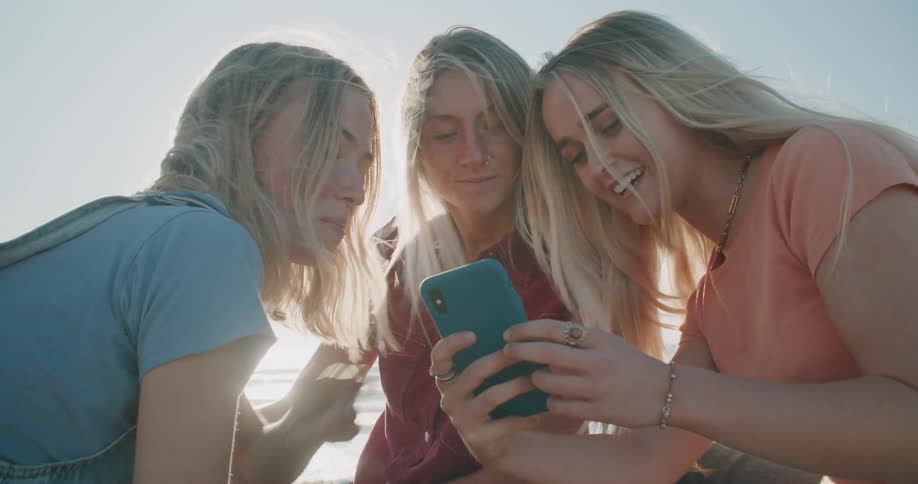 Three women laugh and look at a phone on the beach