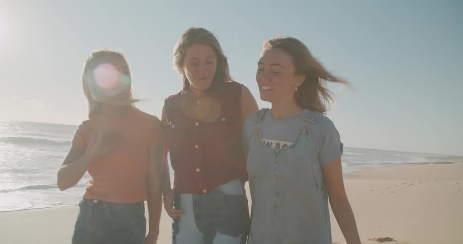 Three young female friends laughing and walking on the white sand beach next to the ocean