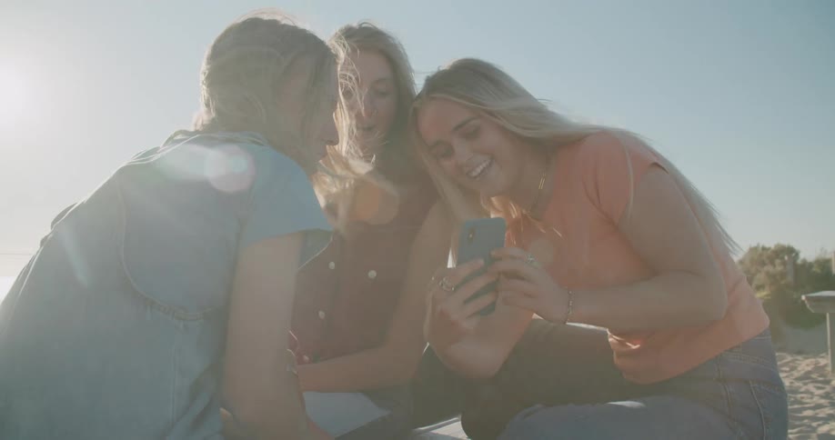 Three teenage girls sit on the shore and laugh and look at a cell phone