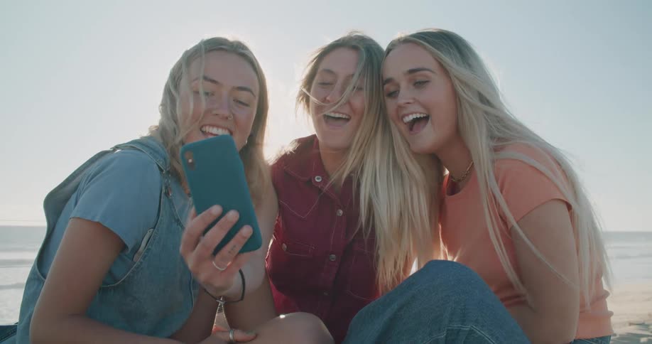 Three blonde teenagers smiling while video chatting on a sunny beach