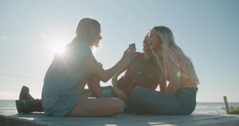 A group of women sitting together on a beach laugh while looking at the screen of a smartphone