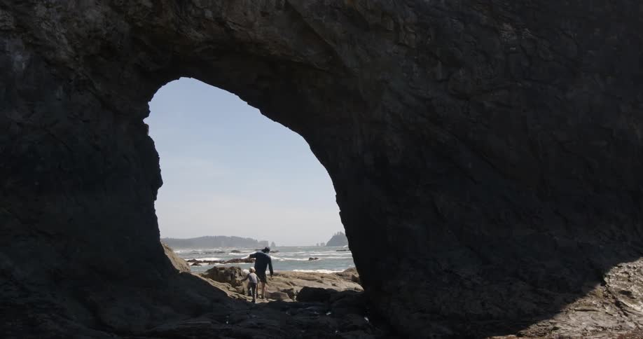 A man and boy walk through a stone arch on a beach at daylight