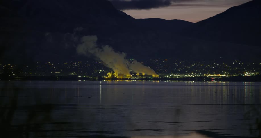 Smoke rising from a factory next to a mountain lake at night