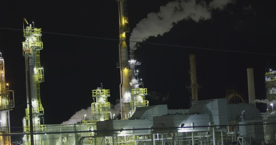 A factory with lights chimneys and smoke with the sky in the background